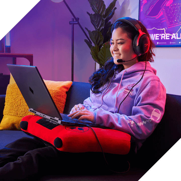 Eine junge Frau mit Gaming-Headset nutzt entspannt einen Laptop auf dem Schoßkissen auf der Couch.