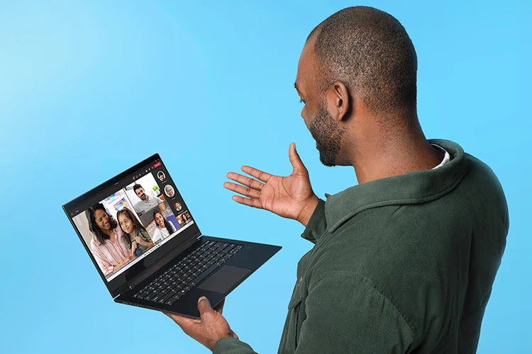 Ein Mann hält einen schwarzen Laptop vor blauem Hintergrund und zeigt lächelnd auf eine Videokonferenz auf dem Display.