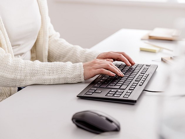Person in hellem Cardigan tippt an einem hellen Schreibtisch auf einer schwarzen Tastatur mit passender Computermaus.