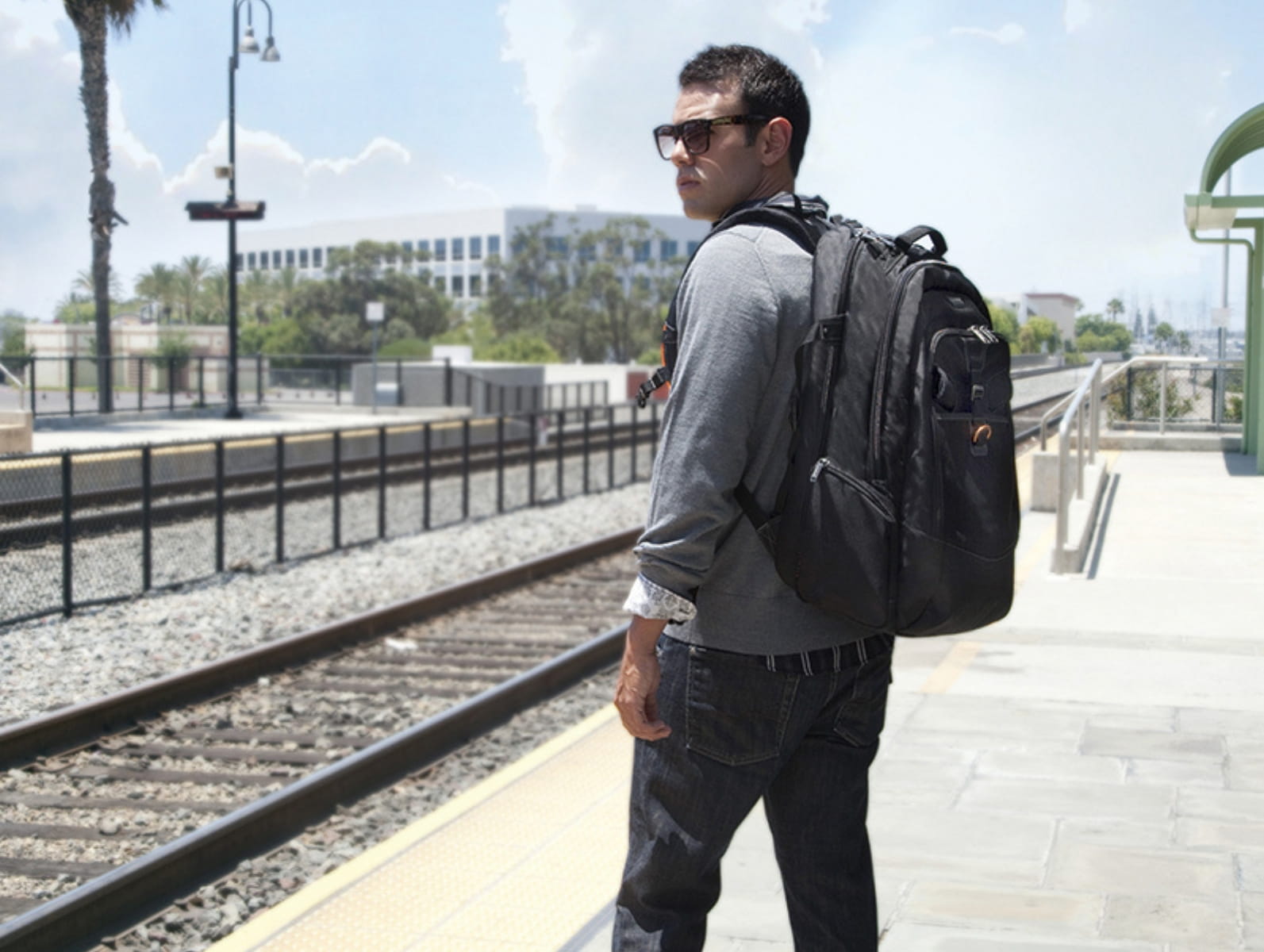 Mann mit grauem Pullover und schwarzem Laptop-Rucksack wartet am sonnigen Bahnsteig