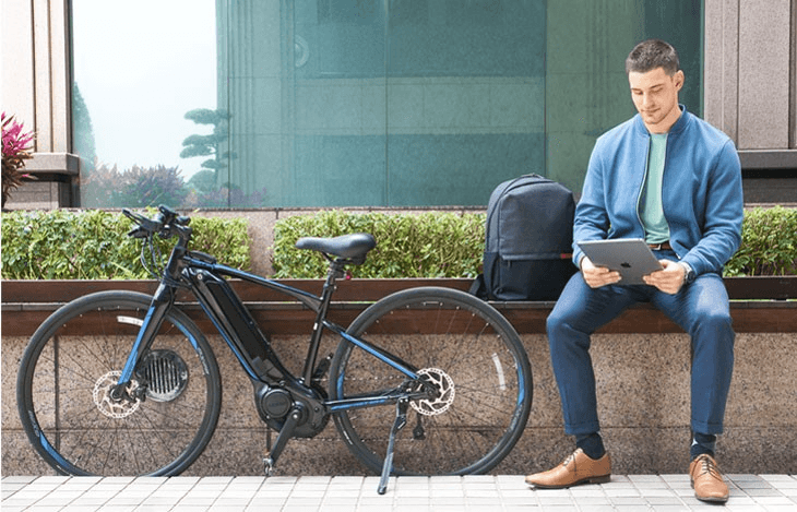 Ein Mann sitzt draußen auf einer Bank, nutzt ein Tablet und blickt auf das Display, daneben stehen ein E-Bike und ein Rucksack.