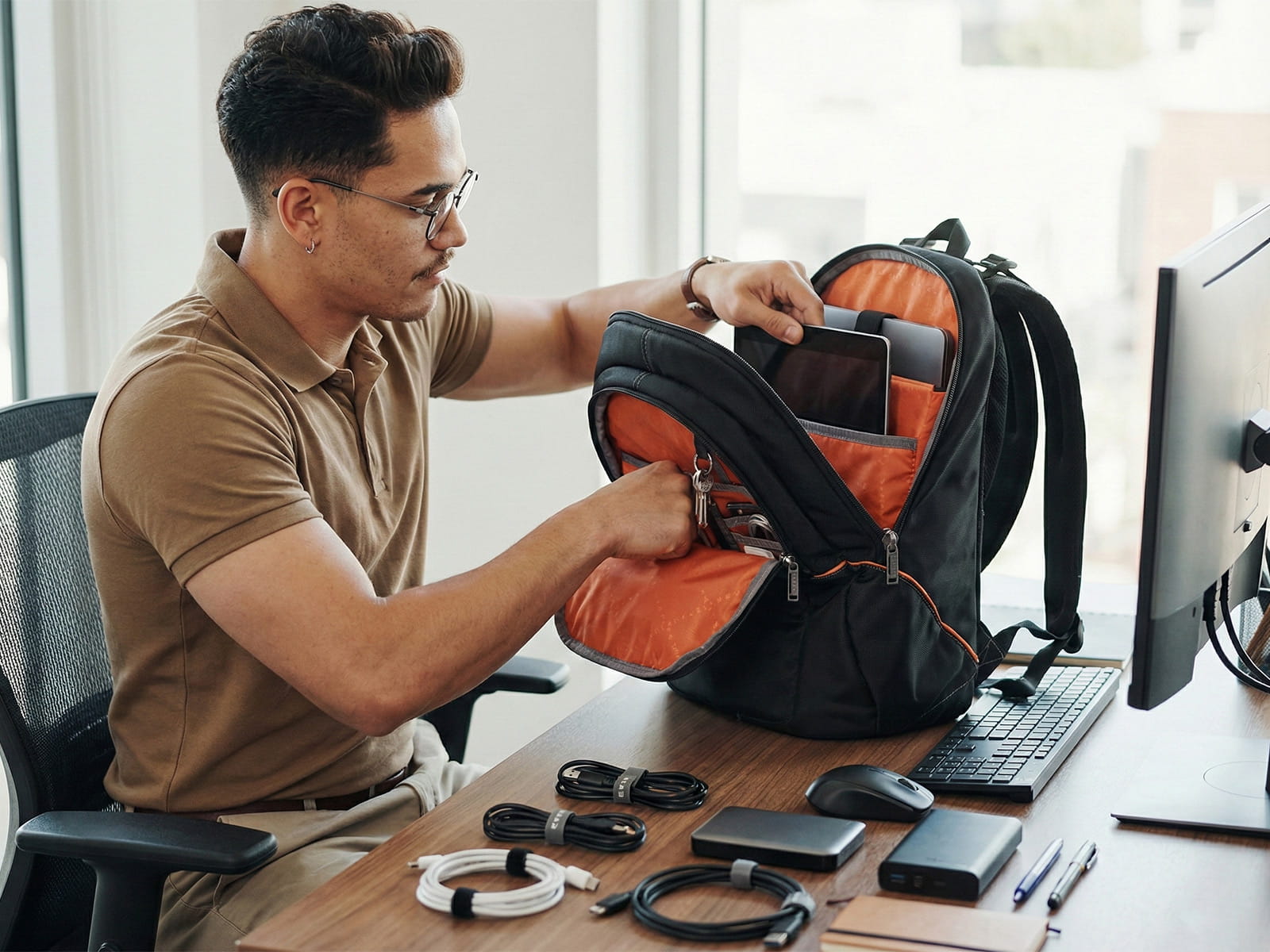 Ein Mann packt am Schreibtisch IT-Zubehör und ein Tablet in einen gut organisierten Laptop-Rucksack