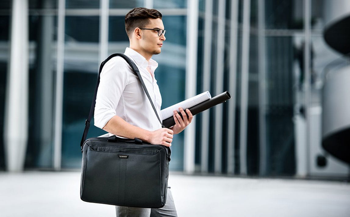 Geschäftssteigernder Business-Mann mit Brille trägt eine schwarze Laptoptasche über der Schulter vor einer modernen Bürofassade.