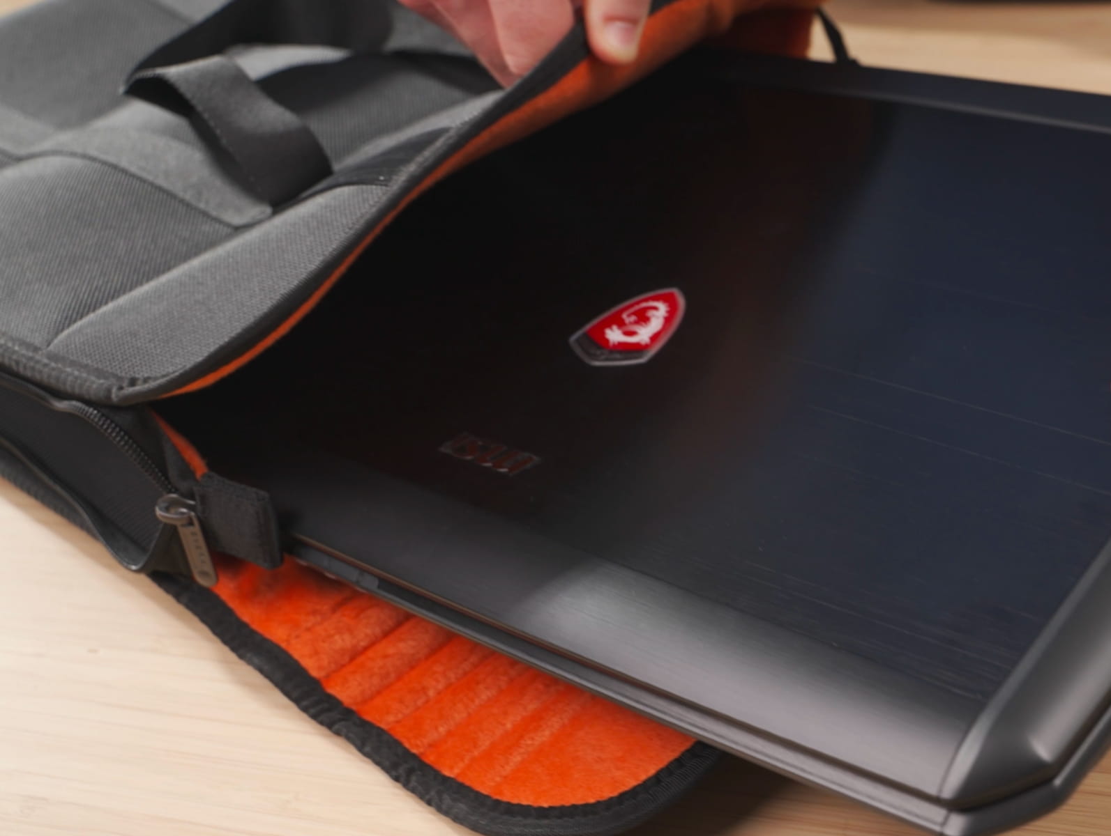 Ein Gaming-Laptop mit rotem MSI-Logo wird in eine gepolsterte, graue Notebooktasche mit orangenem Innenfutter geschoben.