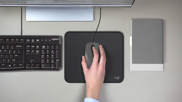 Hand bedient eine graue Logitech Computermaus auf einem Mauspad neben Tastatur und Monitor.