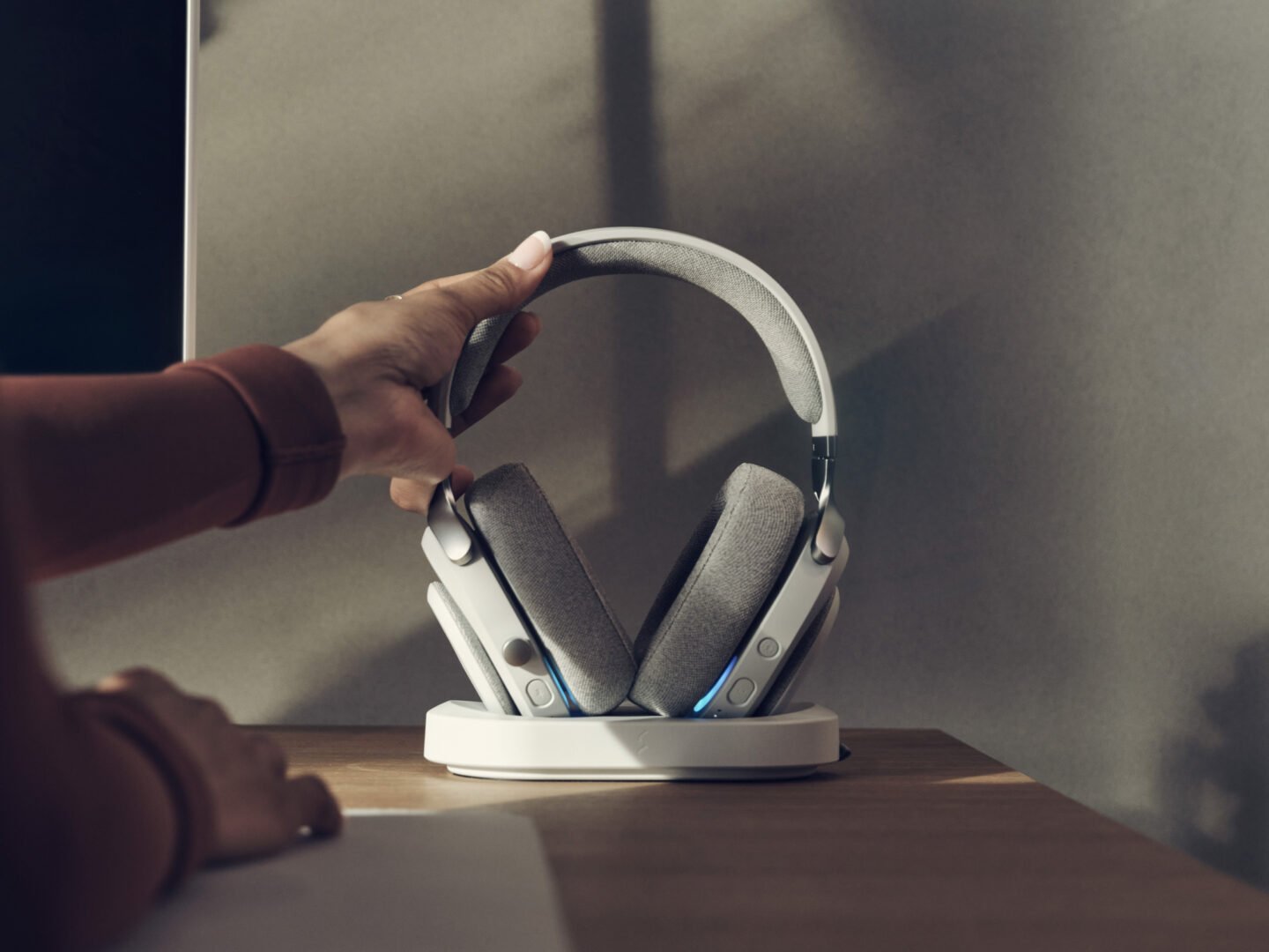 Hand nimmt helle High-End Gaming-Headphones mit Stoffpolstern aus der Ladestation auf dem Schreibtisch.