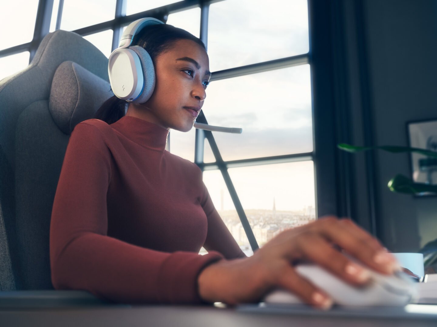 Frau mit Gaming-Headset und Mikrofon sitzt in einem Gaming-Stuhl vor einem Fenster und bedient eine Computer-Maus.