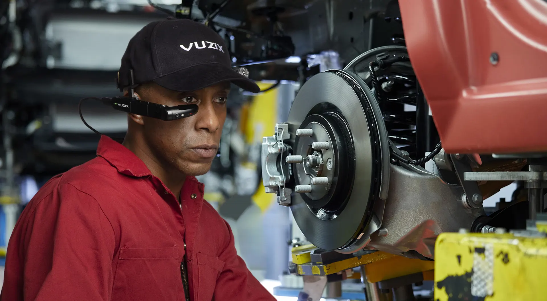 Techniker in rotem Arbeitsanzug nutzt eine Vuzix Smart Glasses VR-Brille bei der Autoreparatur in einer Werkstatt.
