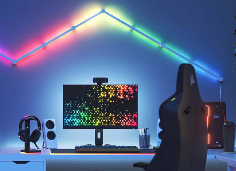 Modernes Gaming-Setup mit Monitor, RGB-Beleuchtung, Gaming-Stuhl und farbigen LED-Wandleisten in einem hellblauen Zimmer