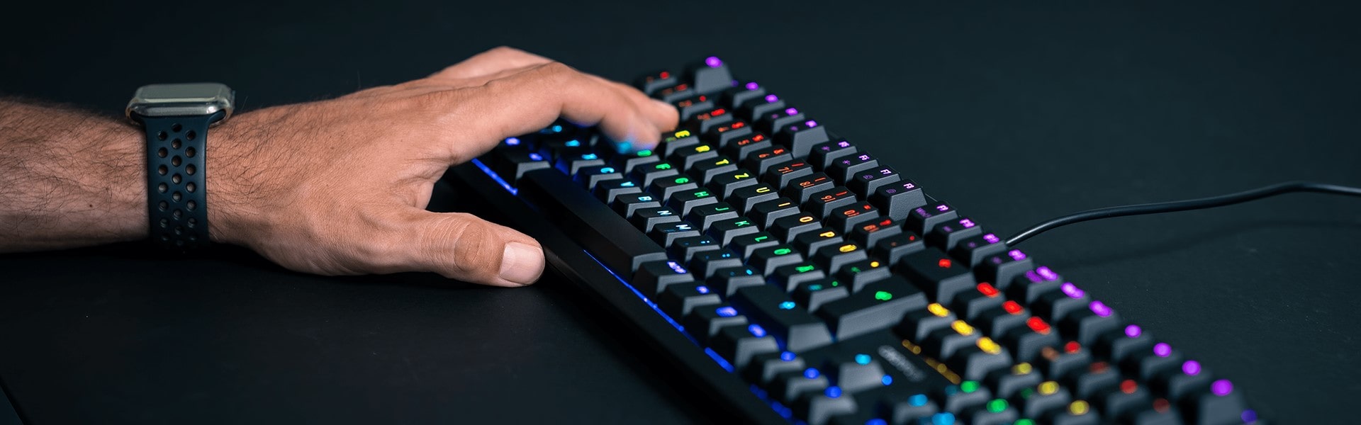 Hand tippt auf eine mechanische Gaming-Tastatur mit bunter RGB-Beleuchtung auf einem dunklen Schreibtisch.