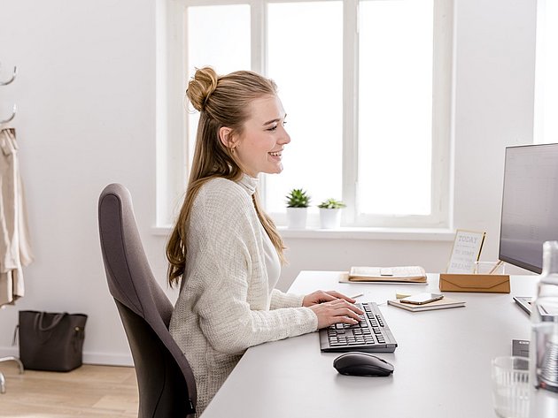 Junge Frau arbeitet an einem ergonomischen Schreibtisch mit Monitor, Tastatur und Maus im hellen Homeoffice