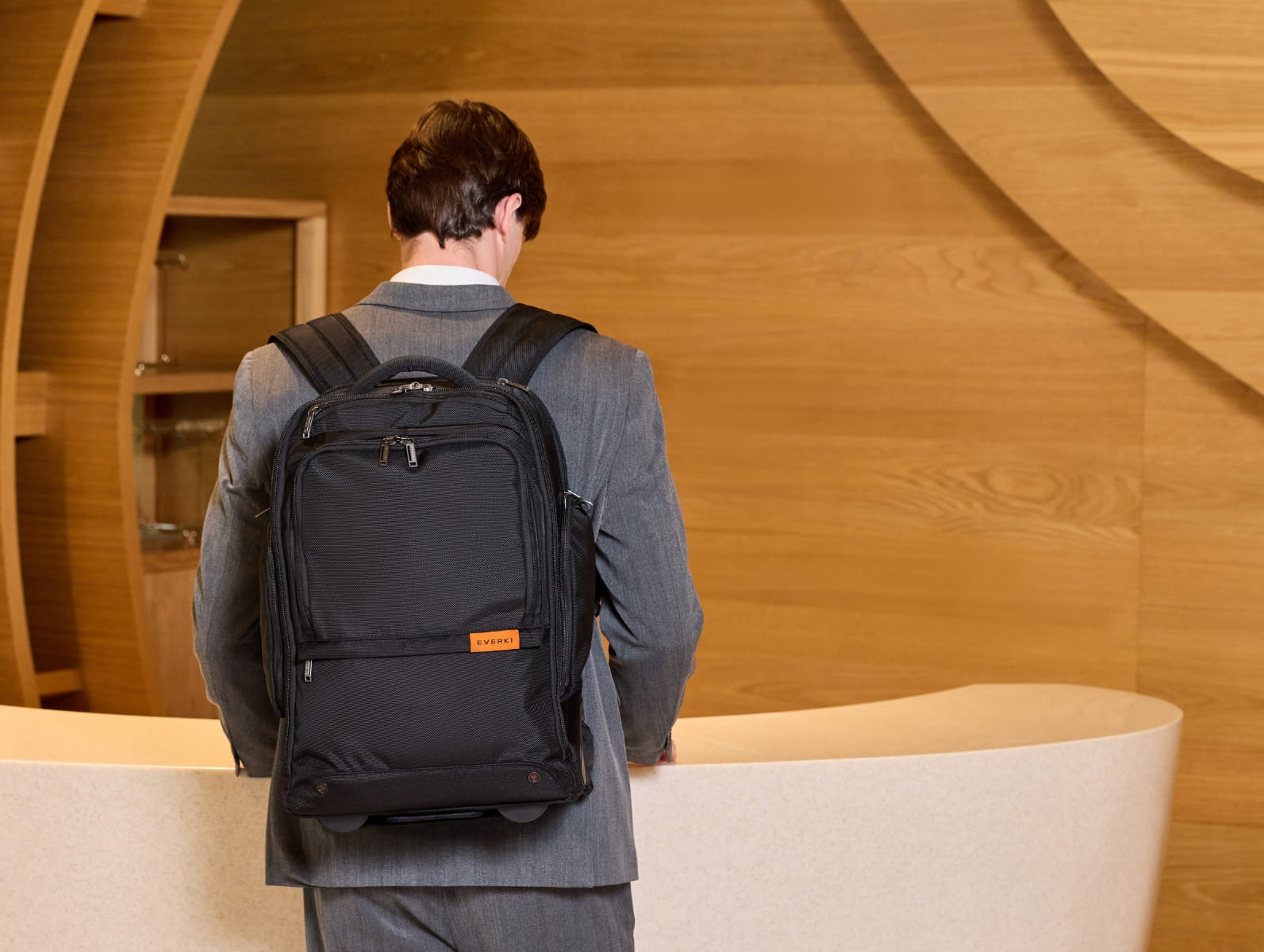 Geschäftsmann in grauem Anzug trägt einen schwarzen Everki Business-Laptop-Rucksack vor einer Holz-Wand.