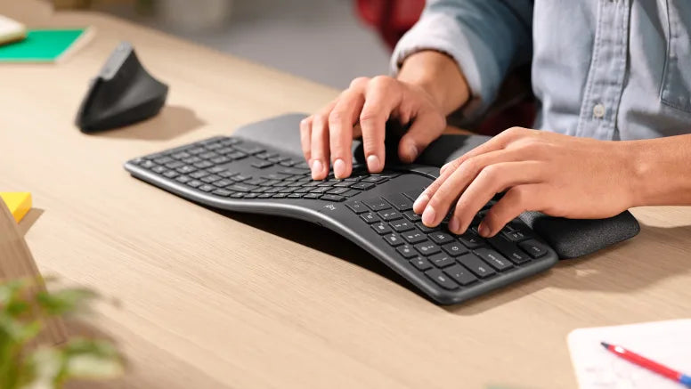 Hände tippen auf einer ergonomischen, geschwungenen kabellosen Tastatur mit Handballenauflage an einem Holztisch.
