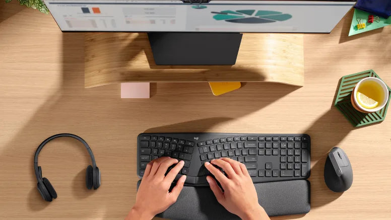 Hände tippen auf ergonomischer Logitech-Tastatur an Schreibtisch mit Monitor, Headset und Vertikalmaus.