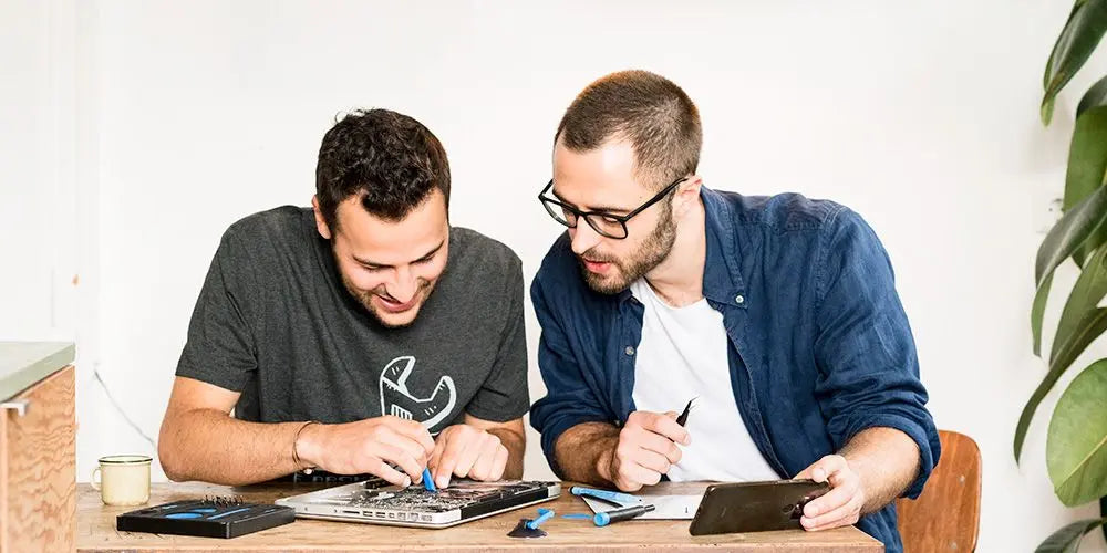 Zwei Techniker reparieren gemeinsam einen Laptop an einem Holztisch mit Werkzeug