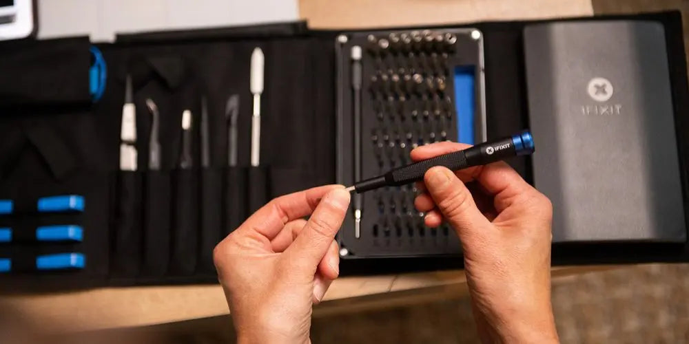 Hände halten einen iFixit-Präzisionsschraubendreher vor einem geöffneten IT-Reparaturset mit Bits und Werkzeugen.