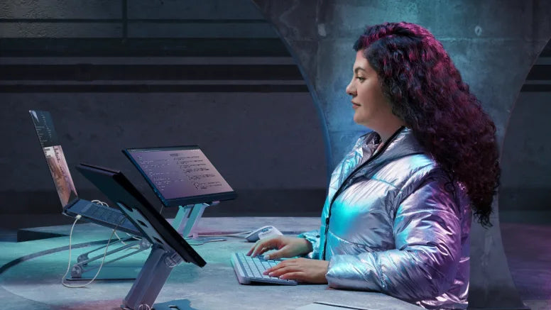Frau in silberner Jacke arbeitet an einem modernen Laptop-Setup mit mehreren Monitoren im futuristischen Design.