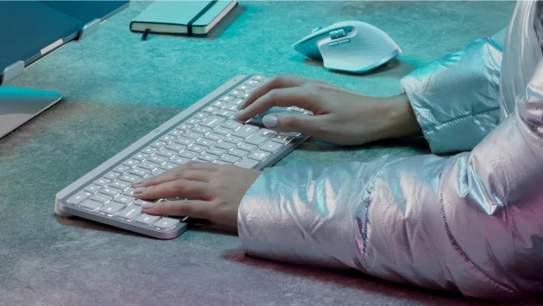 Hände tippen auf einer beleuchteten Wireless-Tastatur neben ergonomischer Computermaus an modernem Schreibtisch.
