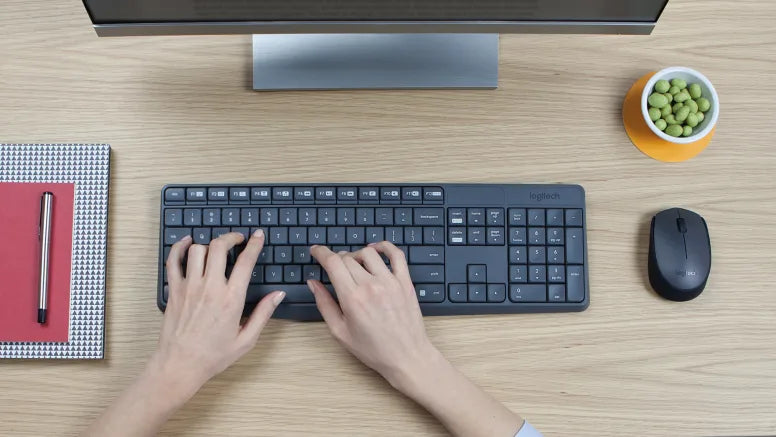 Hände tippen auf einer kabellosen Logitech-Tastatur an einem hellen Holzschreibtisch mit Maus und Monitor.