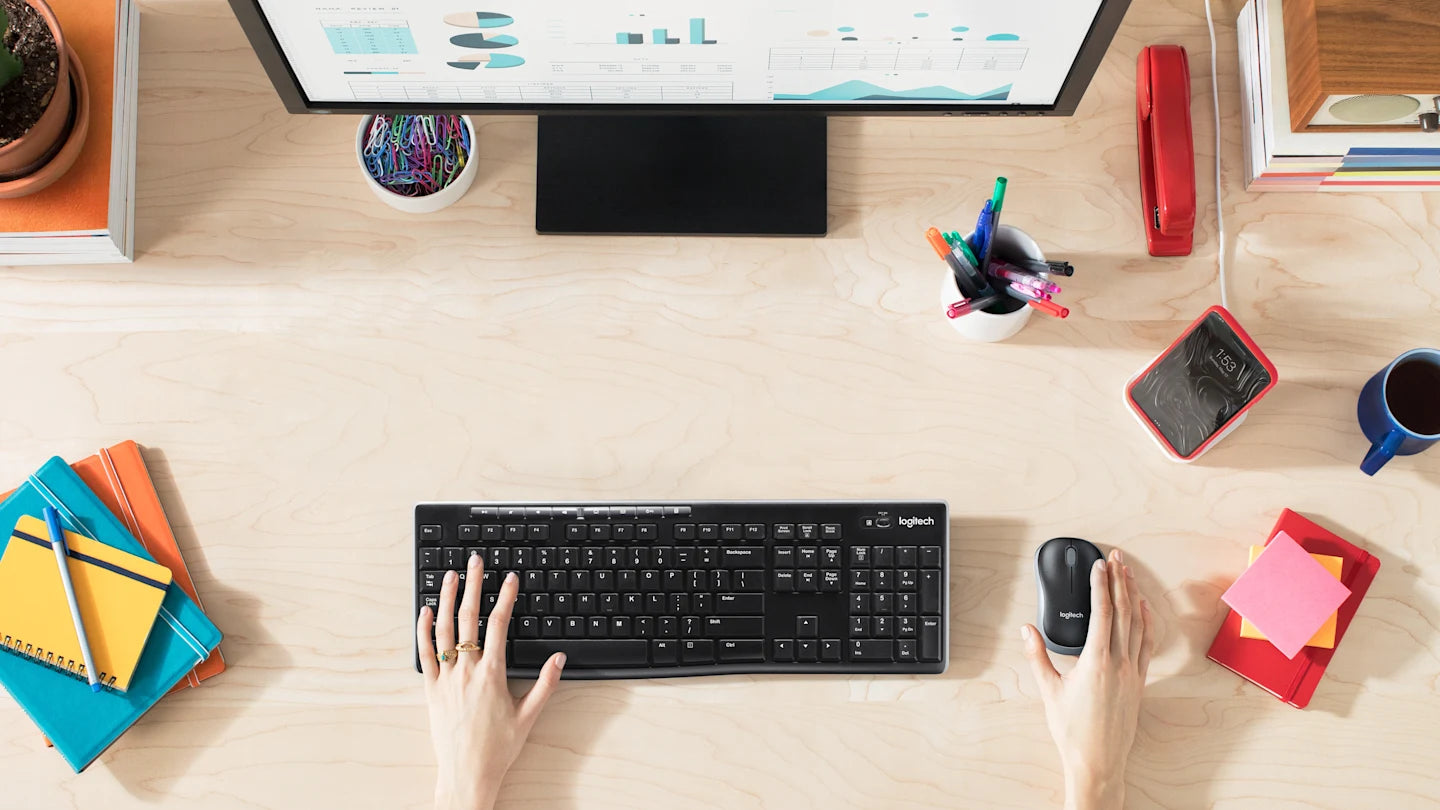 Person arbeitet an einem Holztisch mit schwarzer Logitech Tastatur, Maus, Monitor und Büroaccessoires im Homeoffice.