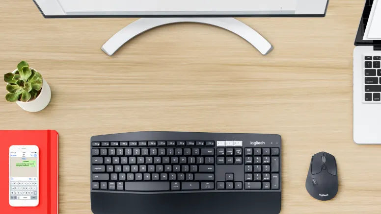 Modernes Homeoffice-Setup mit ergonomischer Logitech Tastatur, Maus, Monitor und Laptop auf Holztisch.