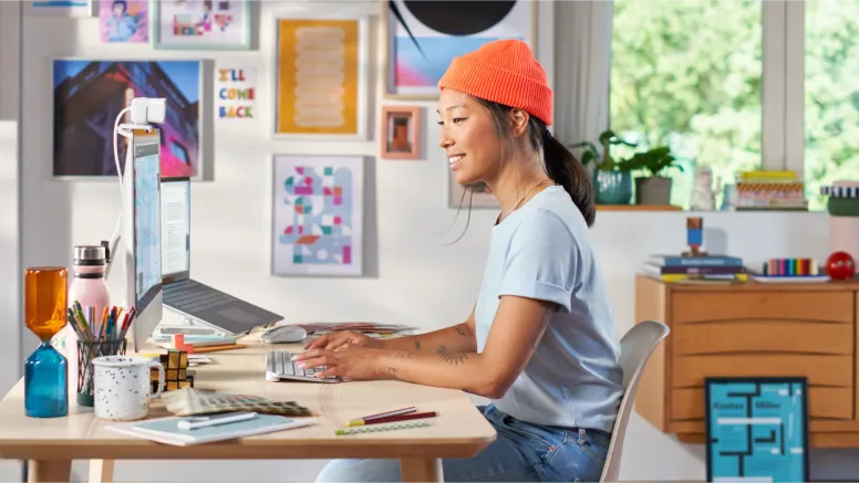 Frau mit orangefarbener Mütze arbeitet an einem modernen Schreibtisch mit Laptop, Monitor und Peripherie.