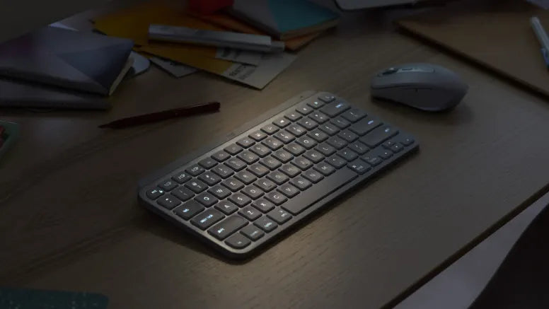 Kompakte kabellose Tastatur mit Hintergrundbeleuchtung und ergonomische Maus auf einem dunklen Holzschreibtisch.